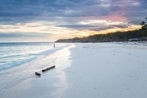 Una playa con aguas tranquilas, una persona caminando y un cielo al atardecer.