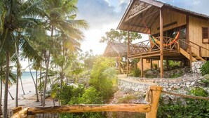 A wooden beach house with a balcony, surrounded by palm trees and a stone wall. - BaraCoco Bungalows (Bira)