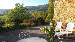 A patio with a round metal table and white chairs, set against a stone wall with a view of a forested hillside.