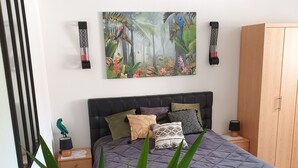 A bedroom with a large bed, a nightstand, a wooden wardrobe, a wall-mounted painting, and a potted plant.