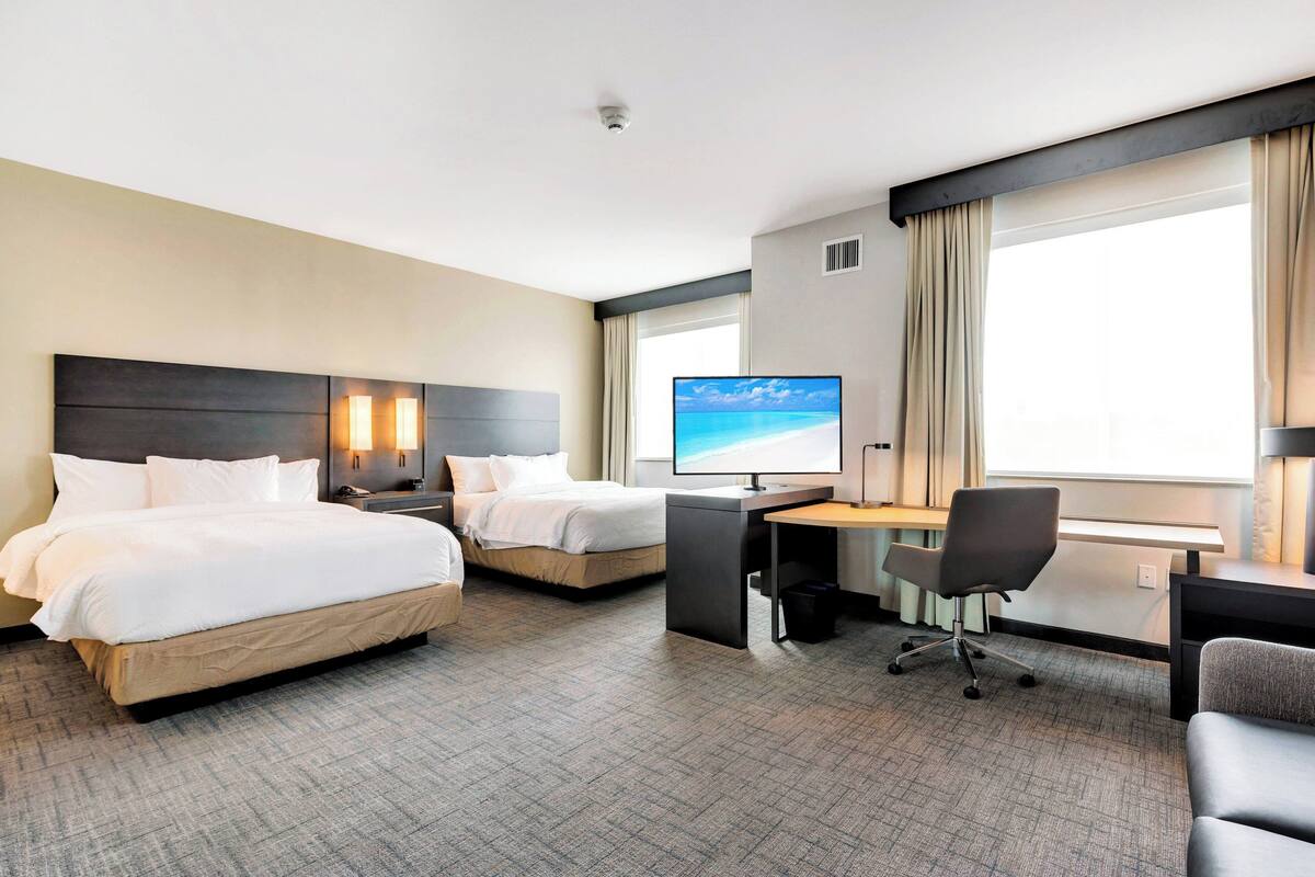 Habitación de hotel con dos camas, un escritorio, una silla y un amplio ventanal con cortinas.