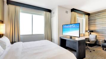 A hotel room with a large bed, a desk with a flat-screen TV, a chair, and a view of the ocean through the window.