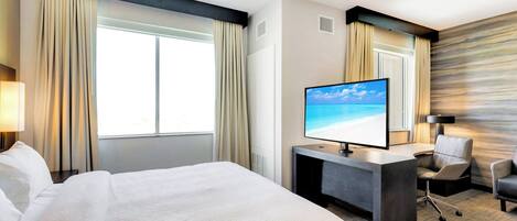 A hotel room with a large bed, a desk with a flat-screen TV, a chair, and a view of the ocean through the window.