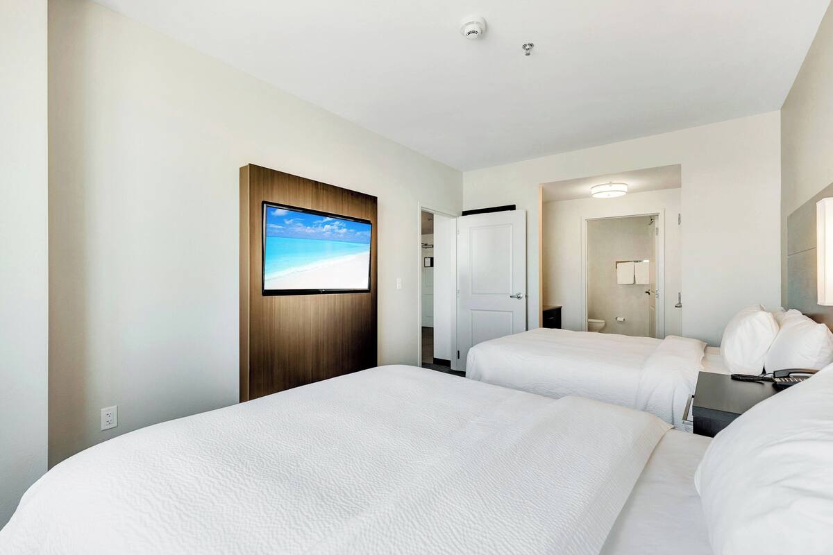 Habitación de hotel con dos camas, un televisor montado en la pared, una puerta que da a un baño y un espejo en la pared opuesta.