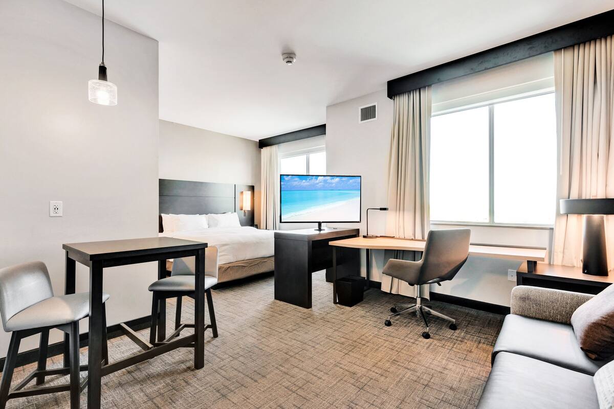Habitación de hotel con cama, escritorio, dos sillas, televisor y ventana con cortinas.