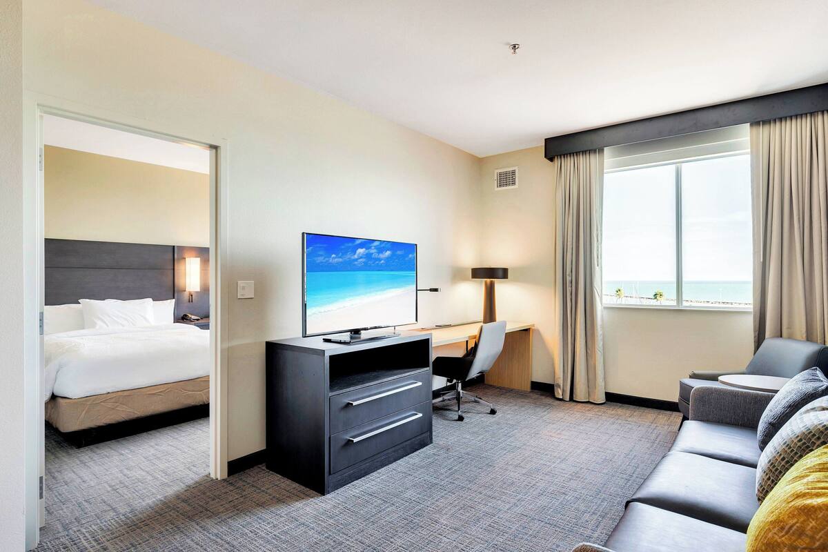 Habitación de hotel con cama, escritorio con computadora, sofá y ventana grande con vista al mar.