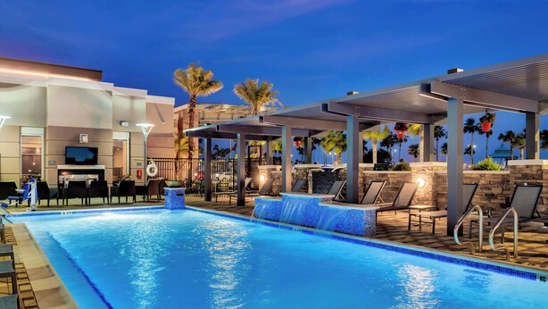 A swimming pool with a waterfall feature, lounge chairs, and a covered seating area with palm trees in the background. - Residence Inn by Marriott Corpus Christi Downtown (Corpus Christi)