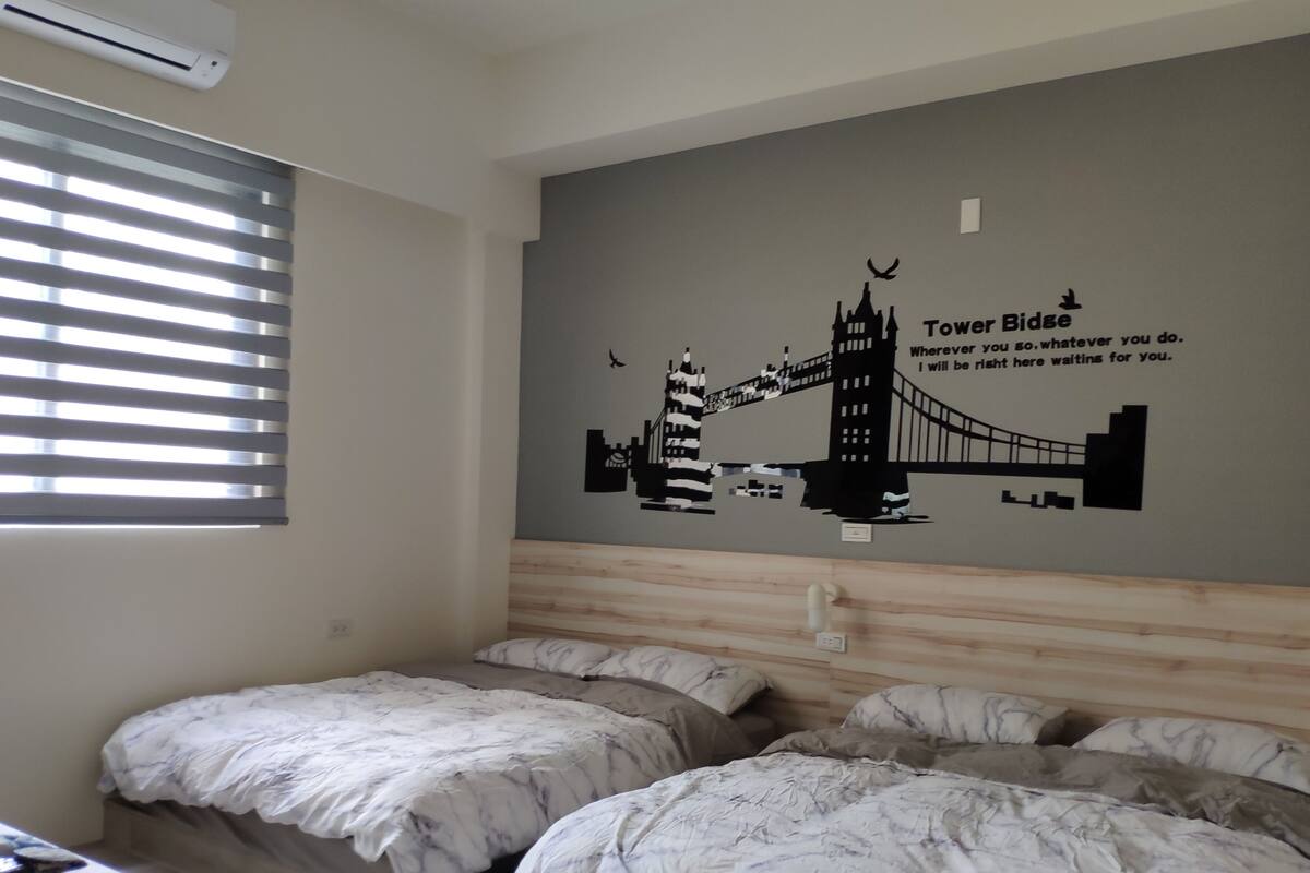 A hotel room with two beds, a large wall mural of the Tower Bridge, and a window with blinds.