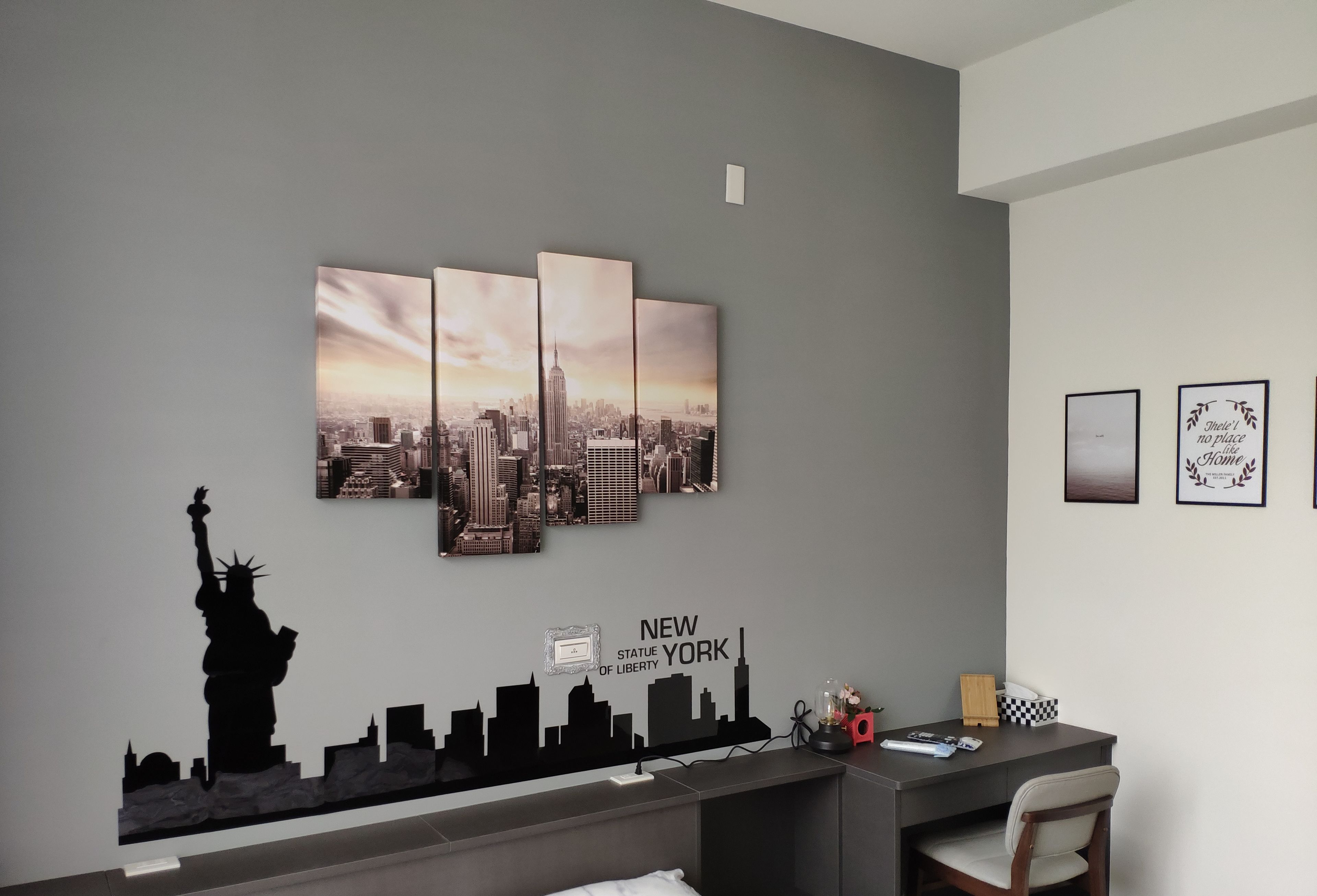 A room with a gray wall, a cityscape artwork, a silhouette of the Statue of Liberty, and a desk with a chair.
