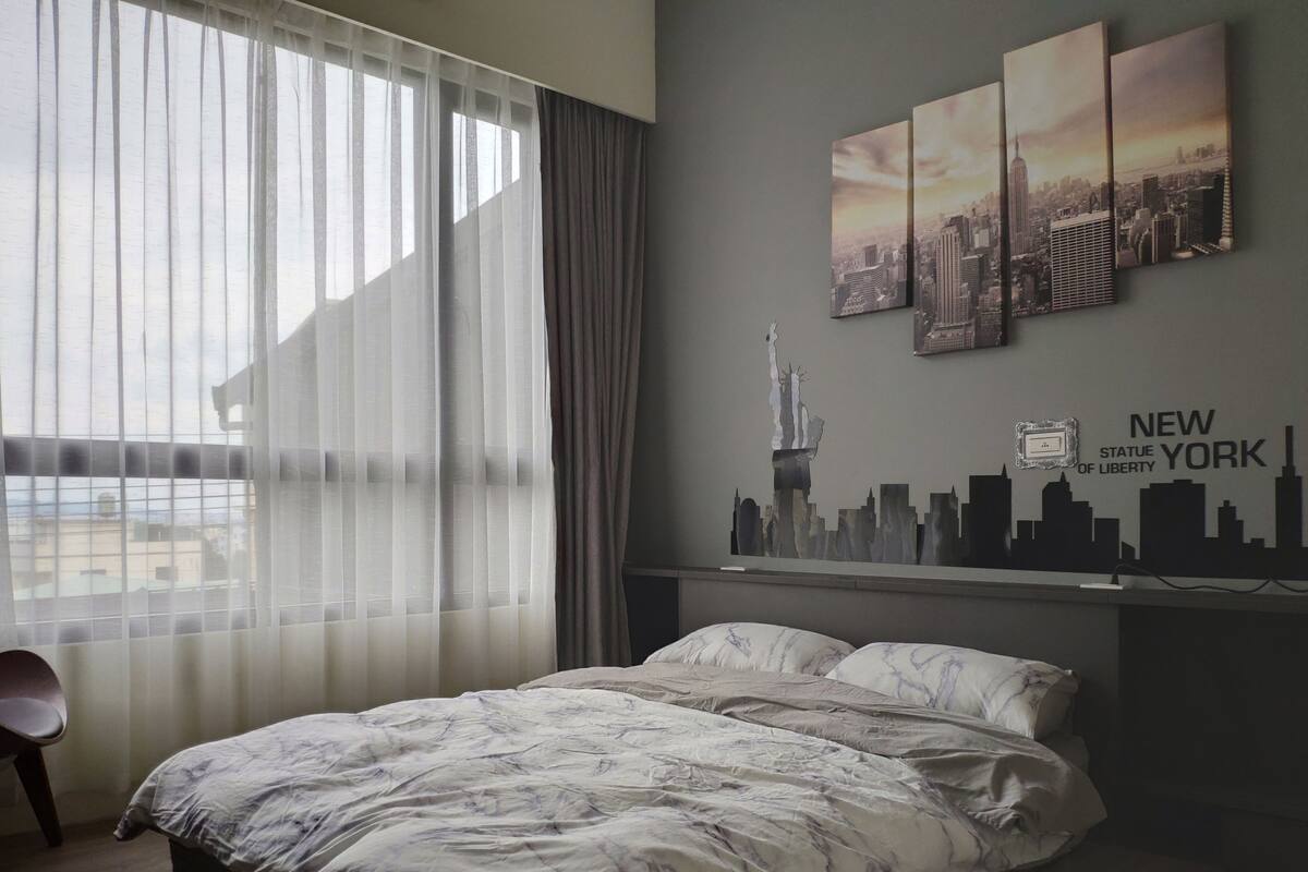 A hotel room with a bed, a chair, a cityscape wall art, and a view of buildings outside.