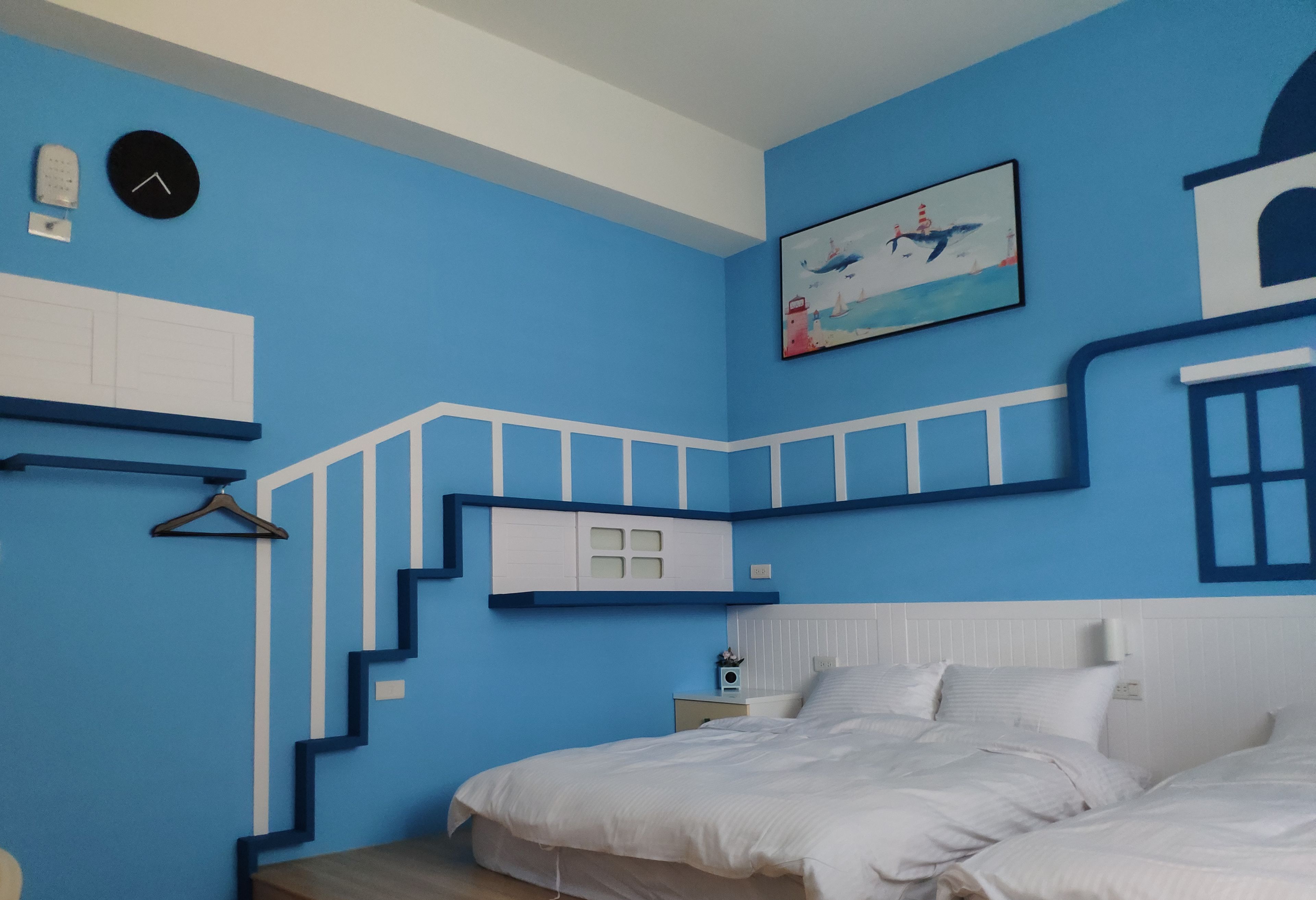 A bedroom with a blue wall, a bed, a nightstand, a clock, and a painting of a lighthouse.