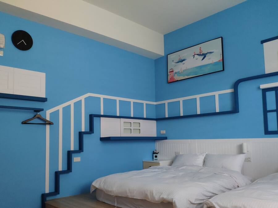 A bedroom with a blue wall, a bed, a nightstand, a clock, and a painting of a lighthouse.
