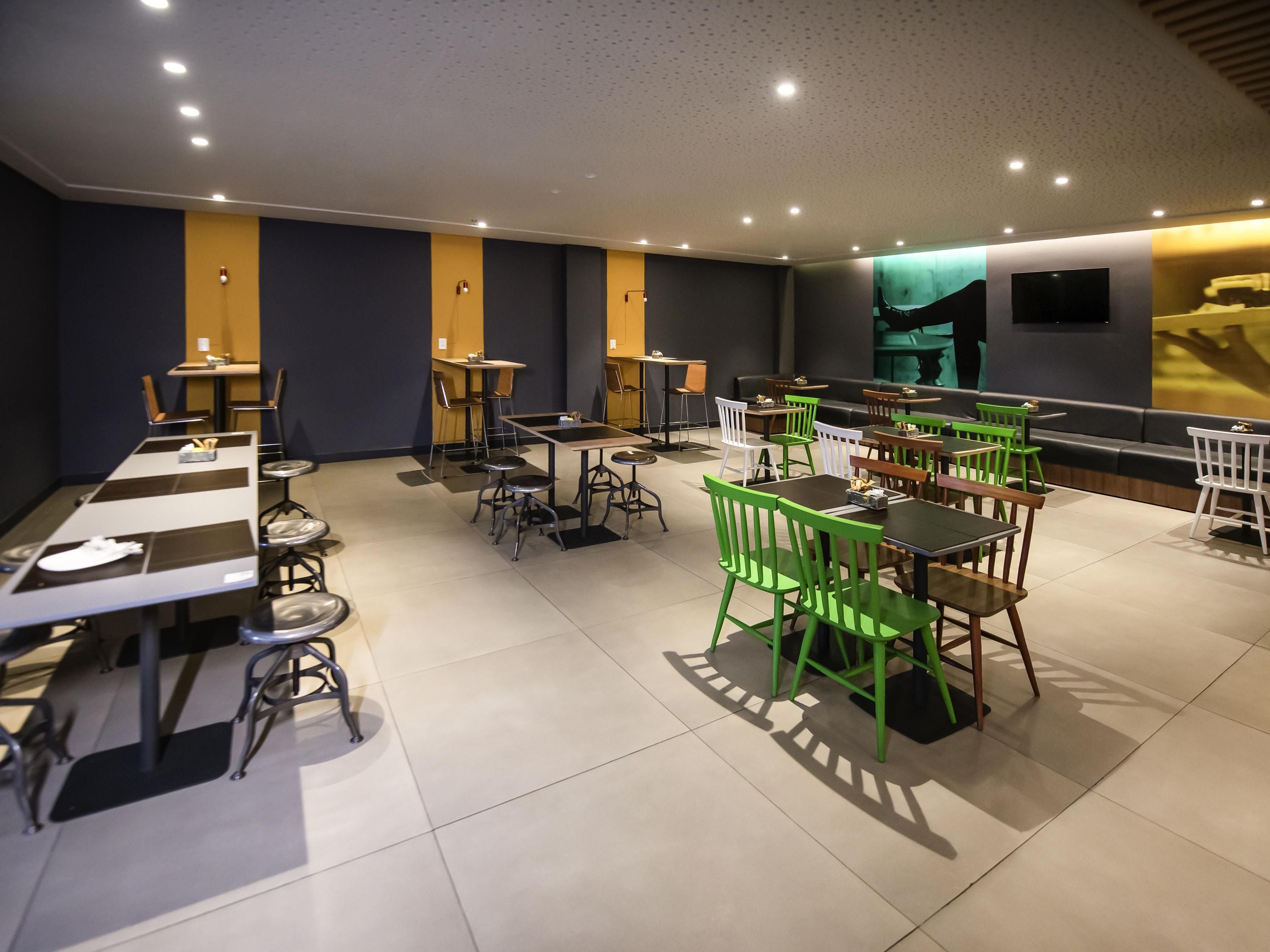 A modern dining area with multiple tables and chairs, a flat-screen TV mounted on the wall, and a sofa in the background.