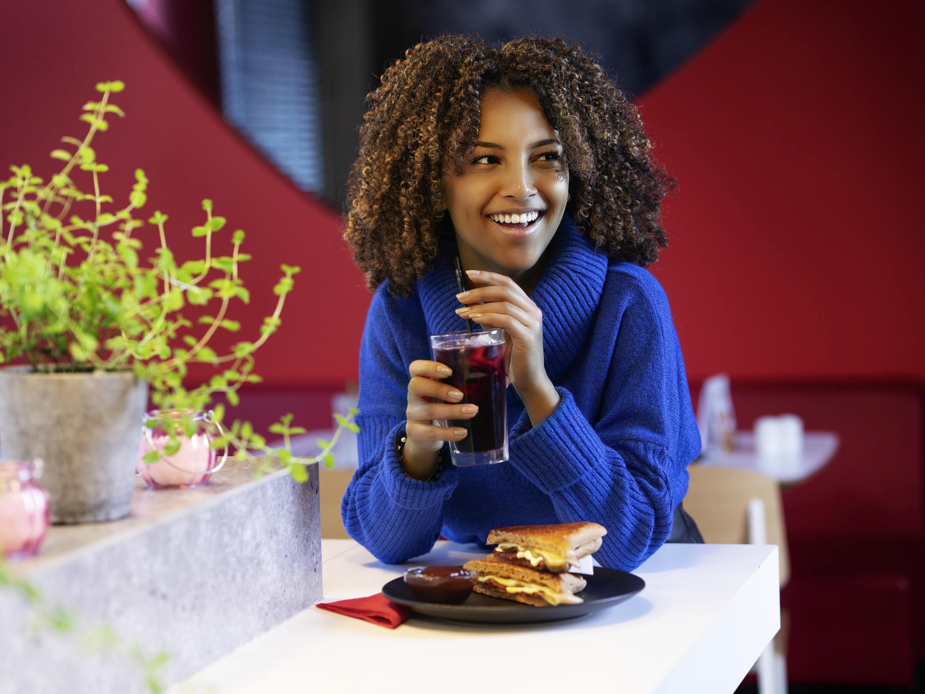 A person sitting at a table with a sandwich and a drink, with a plant and a red wall in the background.