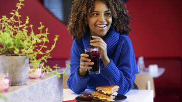 A person sitting at a table with a sandwich and a drink, with a plant and a red wall in the background.