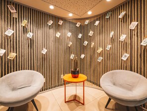 A room with two grey chairs, a small orange table, and a wall decorated with vertical ropes and hanging cards.