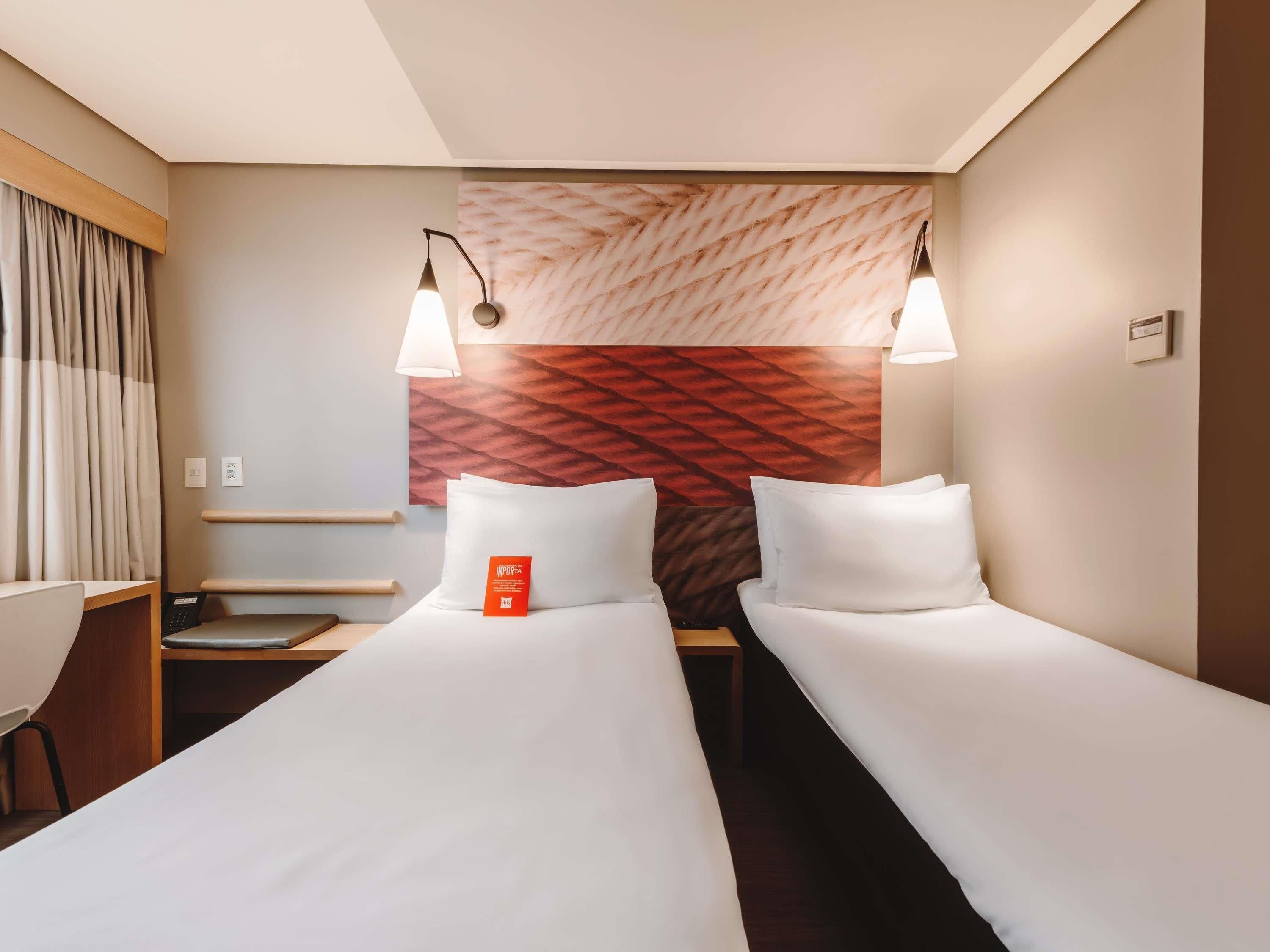 A hotel room with two single beds, a desk, a chair, a wall-mounted lamp, and a patterned headboard.