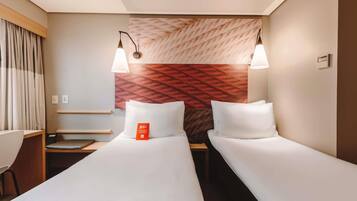 A hotel room with two single beds, a desk, a chair, a wall-mounted lamp, and a patterned headboard.