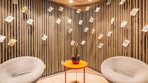 A room with two grey chairs, a small orange table, and a wall decorated with vertical ropes and hanging cards. - Ibis Juazeiro do Norte (Juazeiro do Norte)