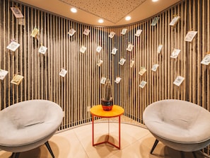 A room with two grey chairs, a small orange table, and a wall decorated with vertical ropes and hanging cards.