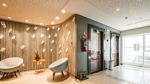 A modern lobby with two grey chairs, a small table with a cactus, a fire extinguisher, and a glass elevator door. - Ibis Juazeiro do Norte (Juazeiro do Norte)
