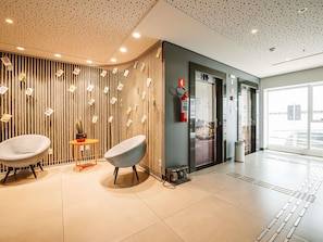 A modern lobby with two grey chairs, a small table with a cactus, a fire extinguisher, and a glass elevator door.