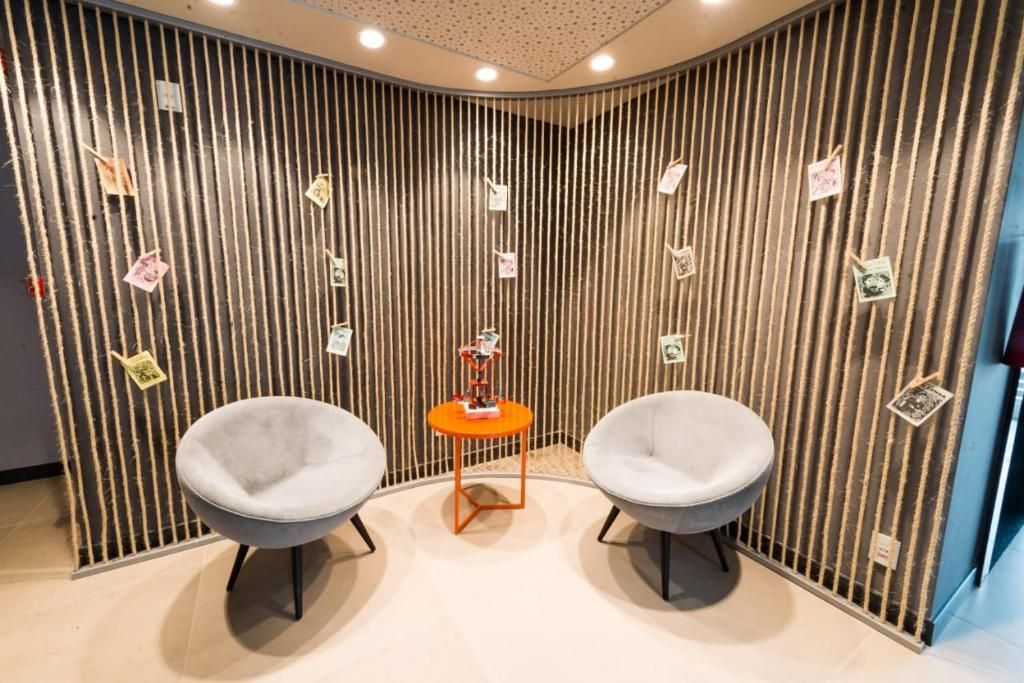 A room with two grey chairs, a small orange table, and a vertical striped wall with various notes.
