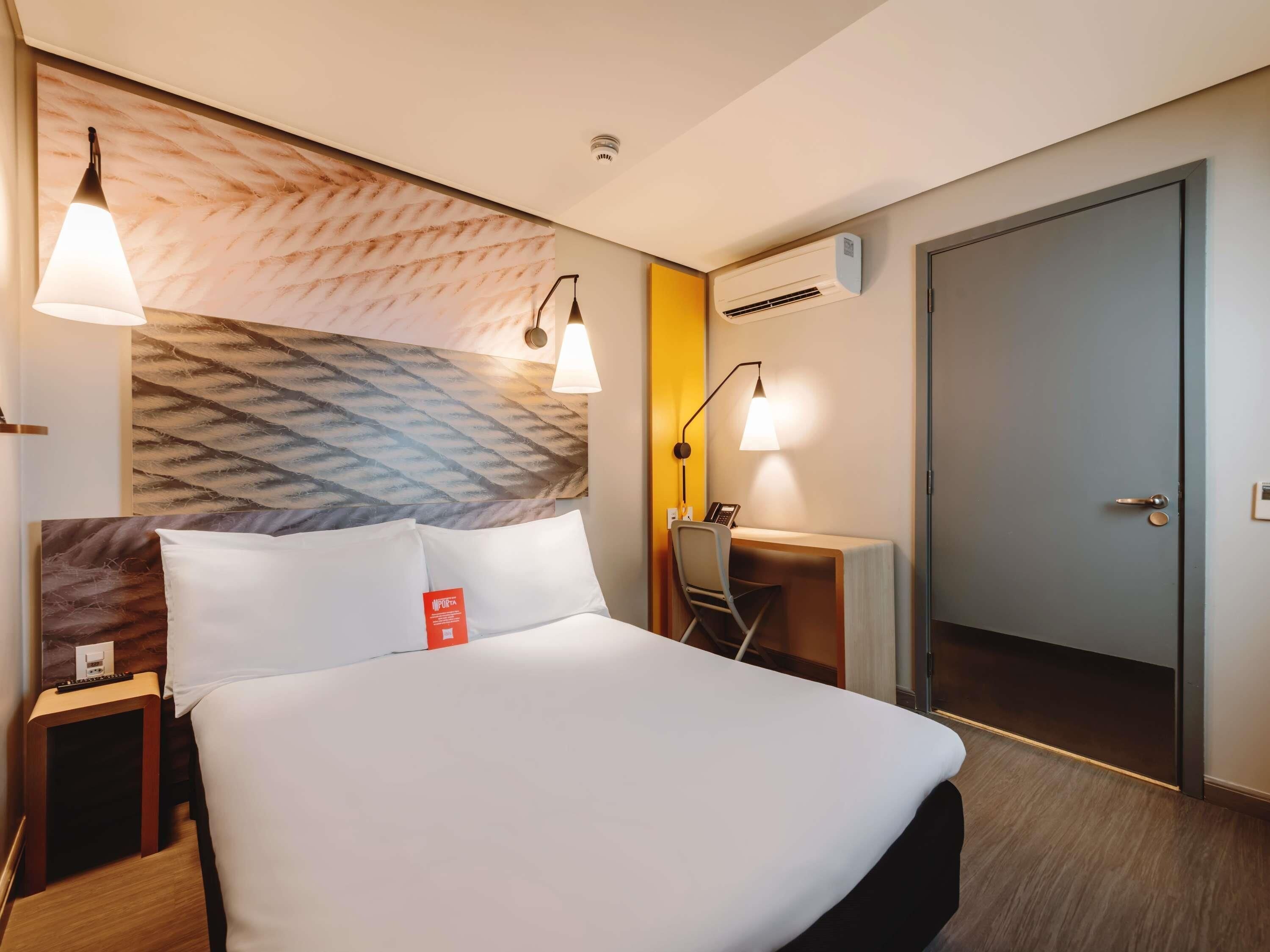 Una habitación de hotel con cama, mesita de noche, escritorio, silla, lámpara y aire acondicionado.