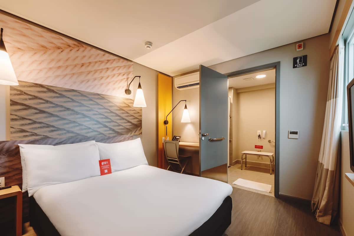 Ein Hotelzimmer mit Bett, Nachttisch, Lampe und einer Tür zu einem Badezimmer.