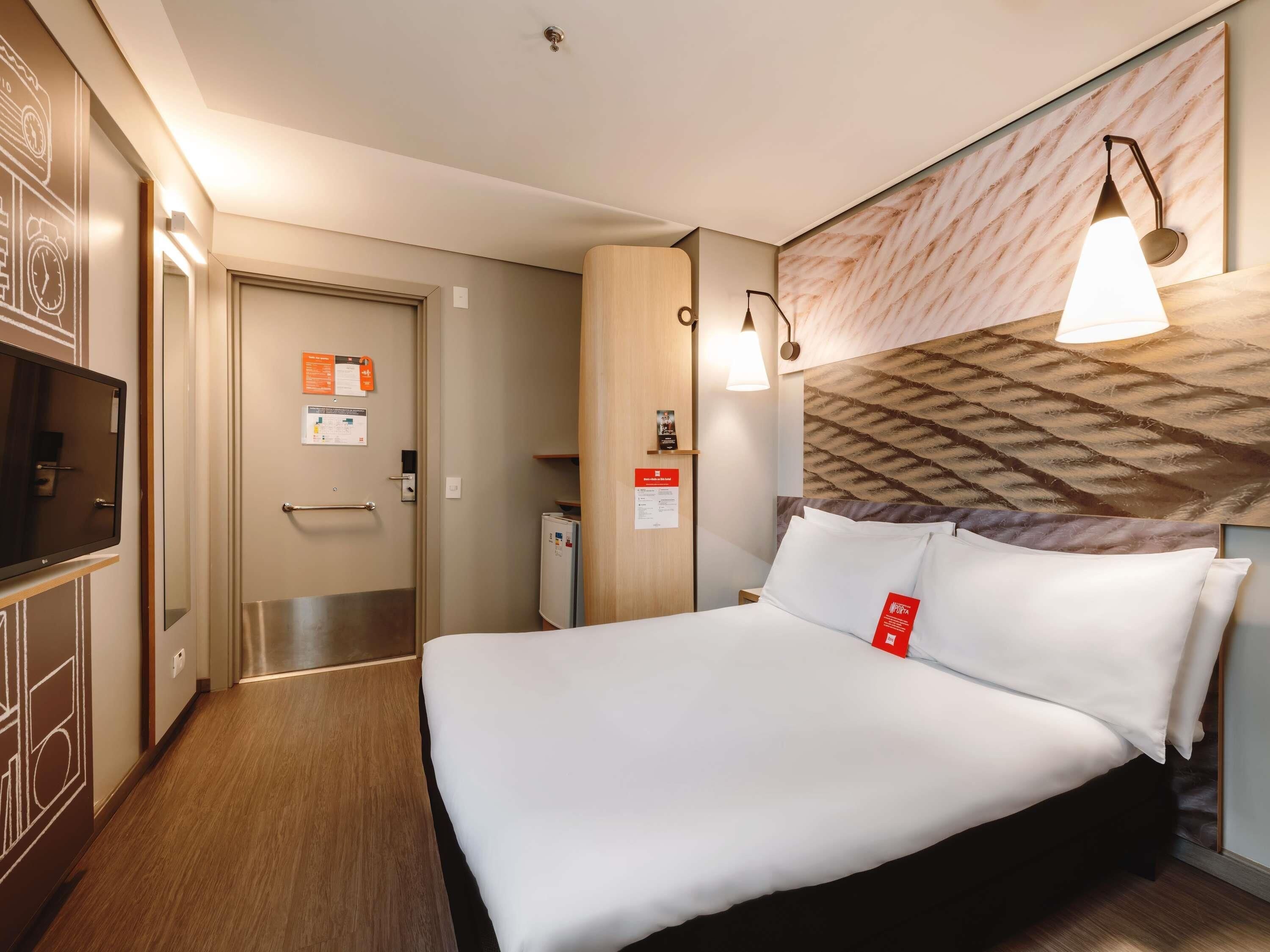 Una habitación de hotel con una cama, televisión, una puerta y una lámpara fijada a la pared.