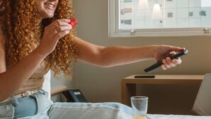 A person sitting on a bed with a remote control, a glass of orange juice, and a plate of food. - Ibis Juazeiro do Norte (Juazeiro do Norte)