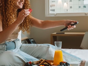 A person sitting on a bed with a remote control, a glass of orange juice, and a plate of food.