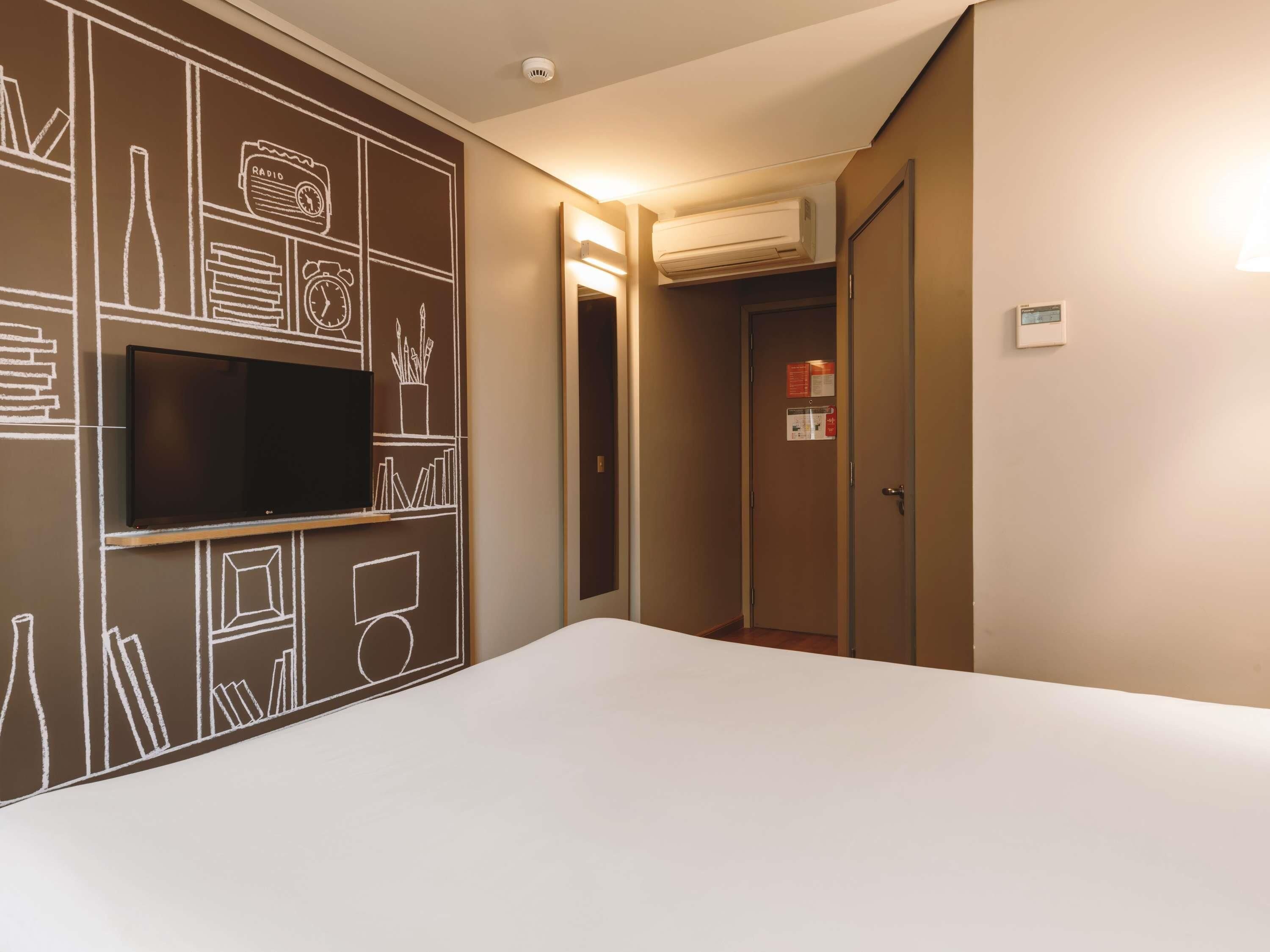 A hotel room with a flat-screen TV mounted on the wall, a bed, an air conditioning unit, and a door with a notice.
