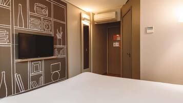 A hotel room with a flat-screen TV mounted on the wall, a bed, an air conditioning unit, and a door with a notice.