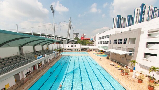 A large indoor swimming pool with multiple lanes, surrounded by a covered seating area and adjacent buildings. - BEAT. Sports Hostel (Singapore)