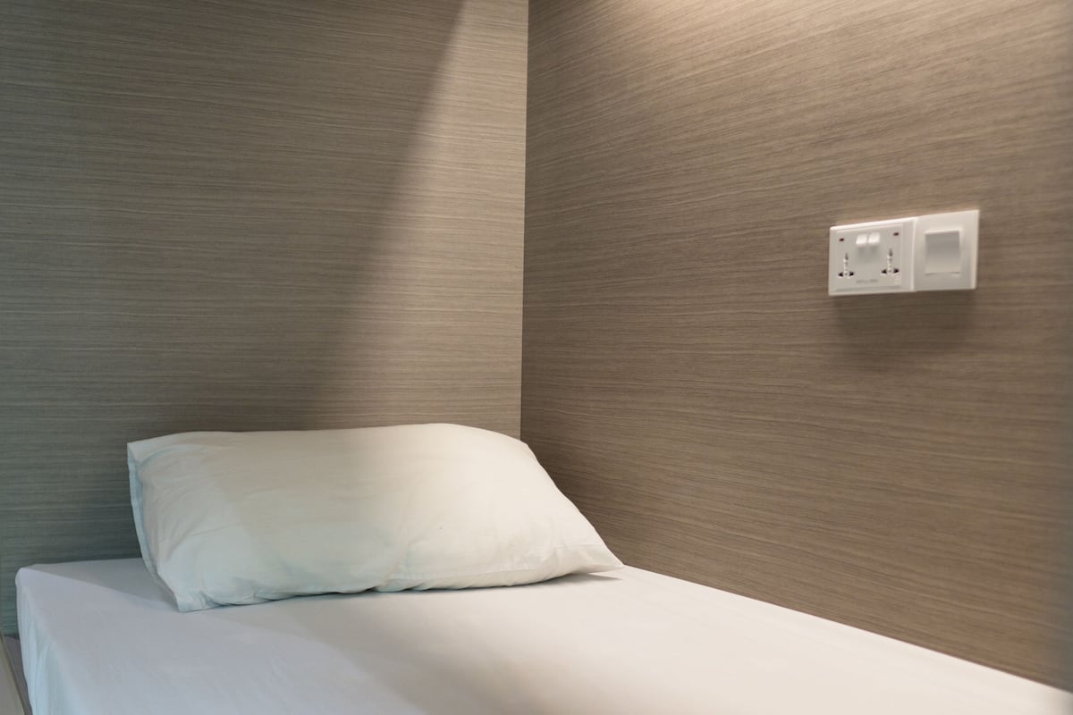 A single bed with a white pillow in a room with wooden panel walls and a wall-mounted electrical outlet.
