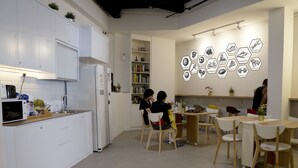 A modern kitchen with white cabinetry, a built-in microwave, and a refrigerator. There are wooden chairs and a table with a vase of flowers. - BEAT. Sports Hostel (Singapore)