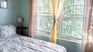Un dormitorio con cama, mesita de noche, ventana con cortinas y vistas al exterior con vegetación.