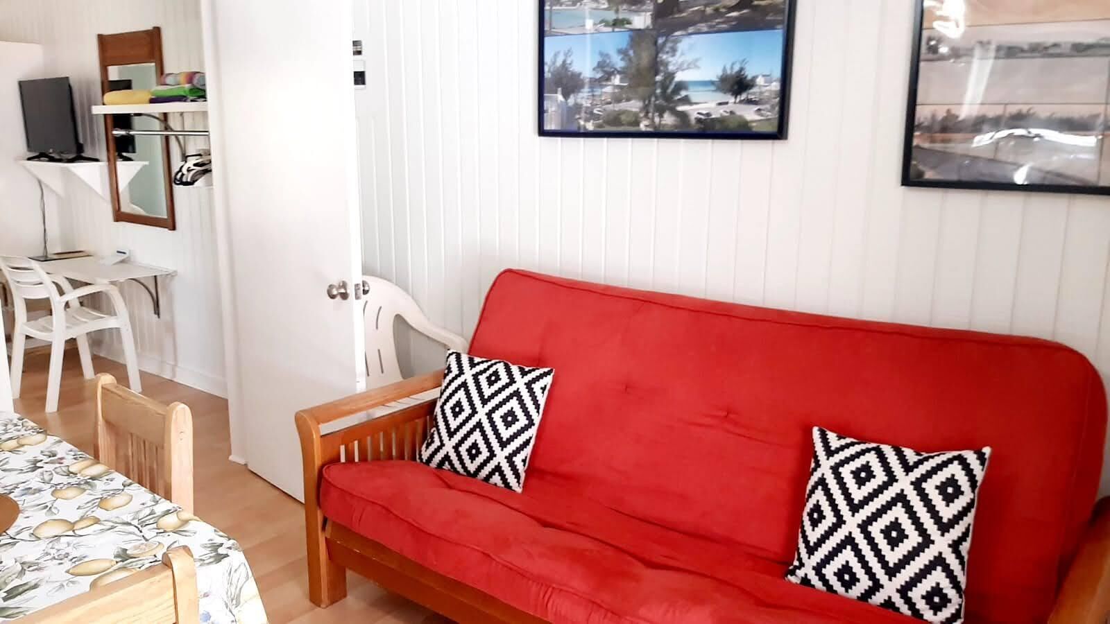 A red sofa with patterned cushions, a wooden chair, a table with a floral tablecloth, and a white wall with a framed picture.