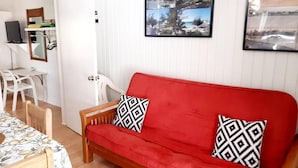 Un sofá rojo con cojines estampados, una silla de madera, una mesa con mantel floral y una pared blanca con un cuadro enmarcado.