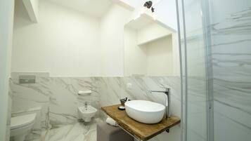 Un baño moderno con piso de mármol, tocador de madera, un lavamanos grande, un bidé y una cabina de ducha de vidrio.