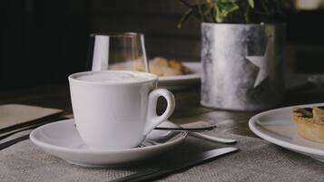 Uma chávena de café branco num pires com uma colher, um copo de água e um jarro de metal sobre uma mesa.