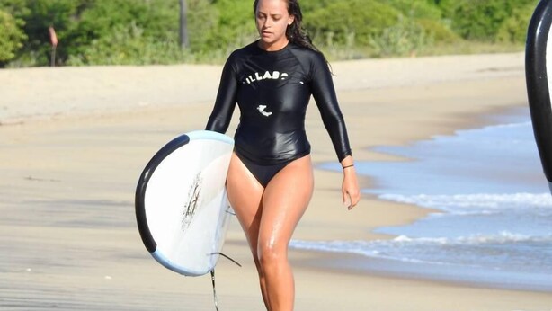 A person in a black swimsuit carrying a surfboard on a sandy beach with greenery in the background. - Exotic Beach Hotel (Pottuvil)