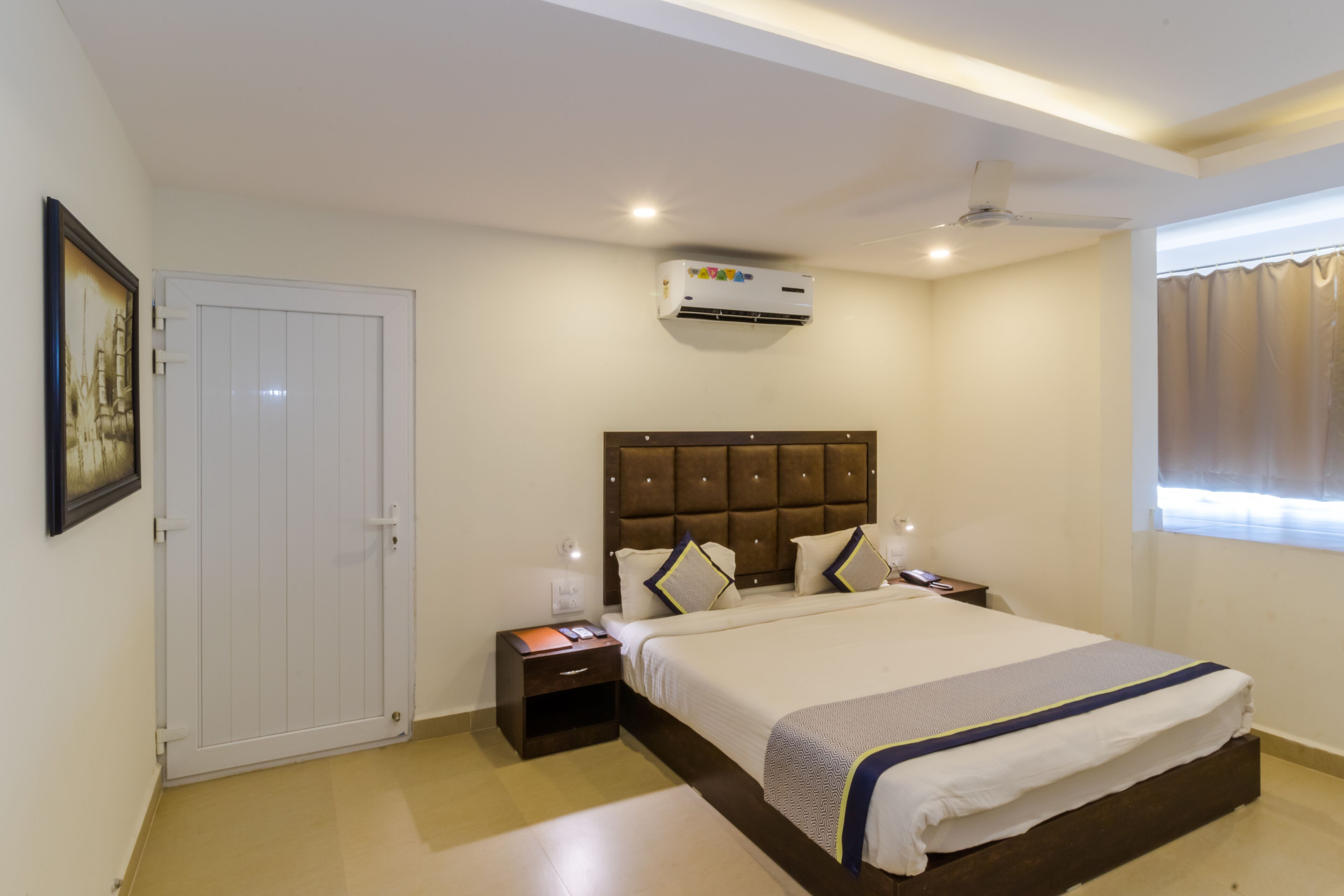 Une chambre à coucher comprenant un lit, des tables de chevet, un appareil de climatisation, un ventilateur de plafond et une porte.