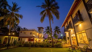 A two-story building with a balcony, surrounded by palm trees and a well-lit garden at dusk. - La Vaiencia Beach Resort (Morjim)