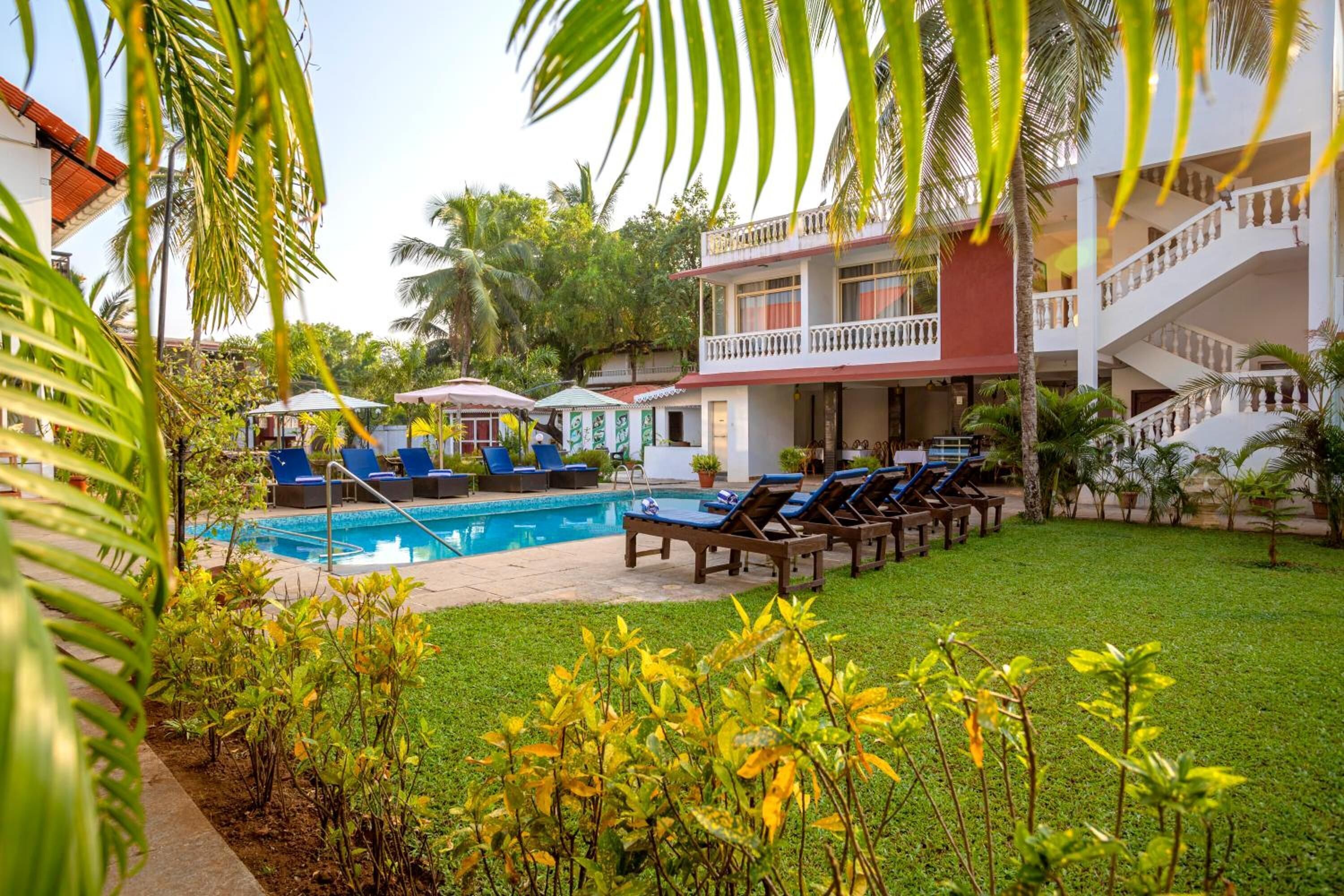 Zona de piscina con sillones reclinables y sombrillas, rodeada de follaje y un edificio de varios pisos con balcones.