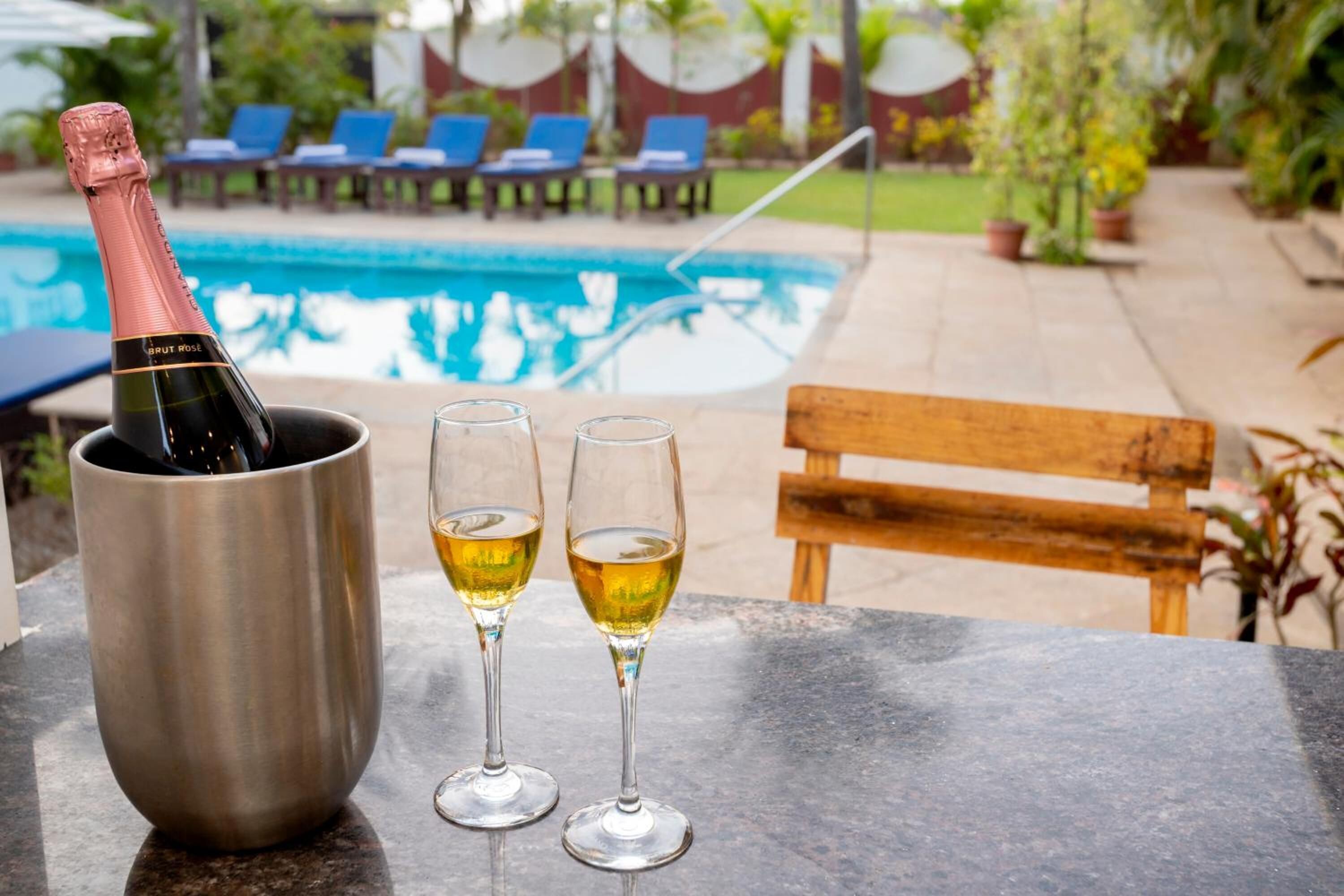 A bottle of champagne in a metal bucket, two glasses of champagne, a pool, and a wooden bench.