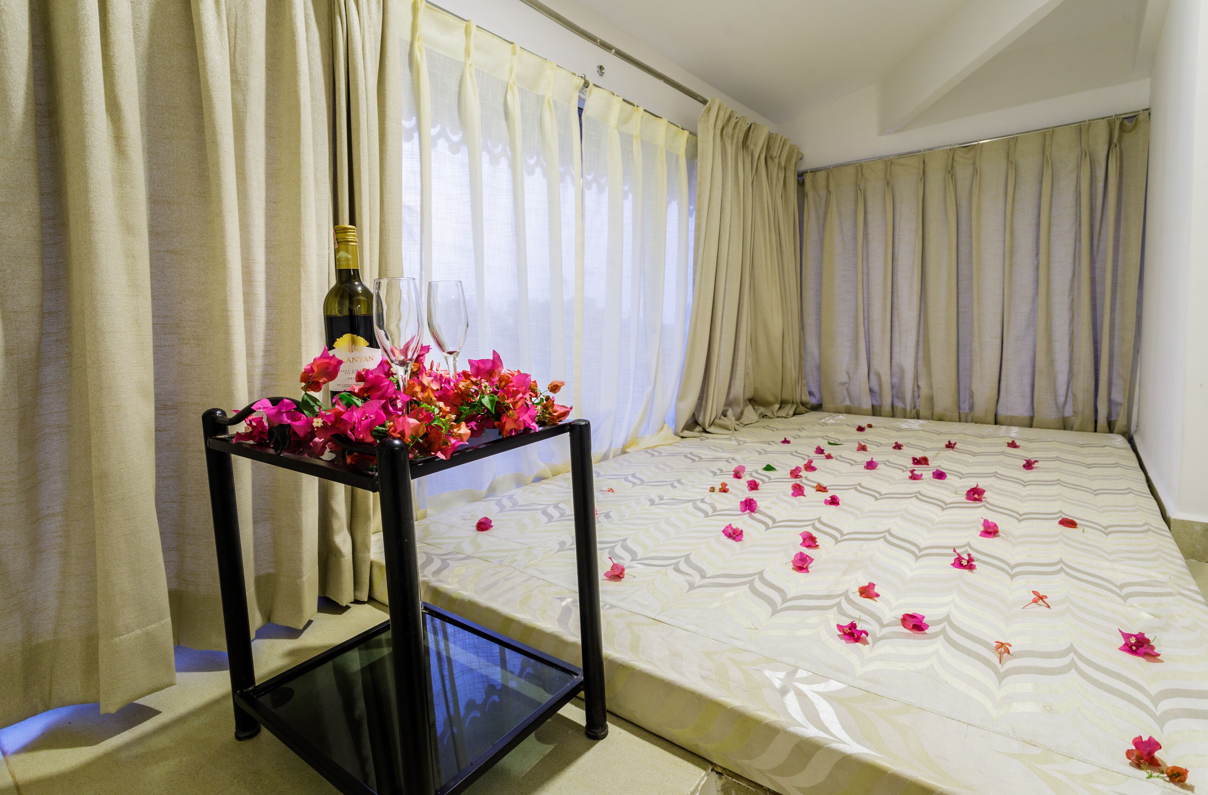 A hotel room with a bed, curtains, a small table with a bottle and flowers, and scattered petals on the bed.