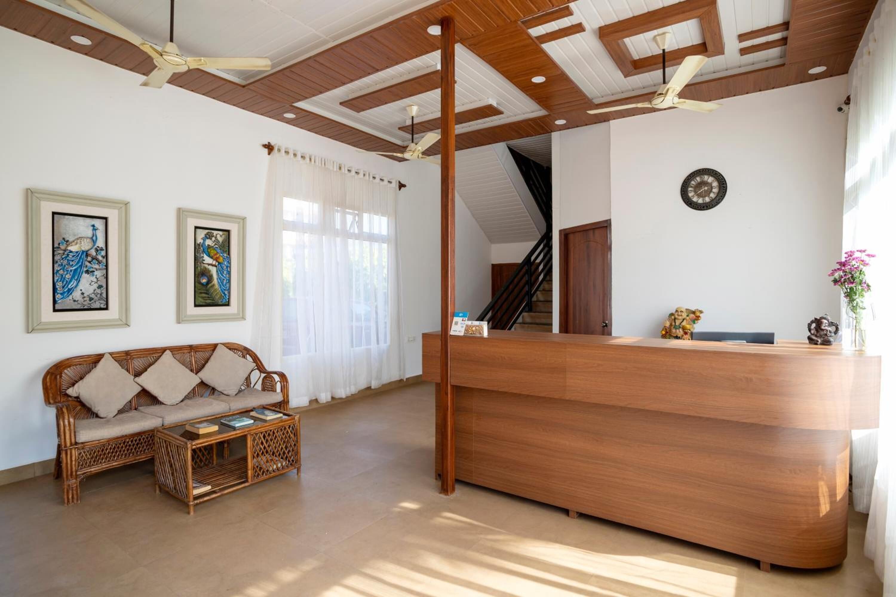 A reception area with a wooden reception desk, a sofa, and a coffee table. There are ceiling fans, a clock, and framed artwork on the walls.