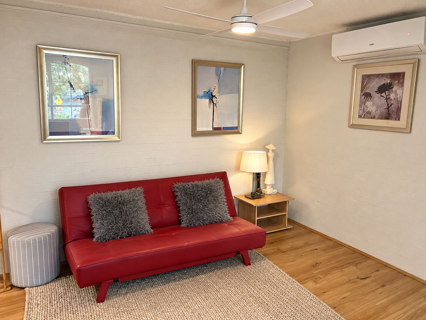 A red sofa with grey cushions, a small side table with a lamp, and framed artwork on the walls.